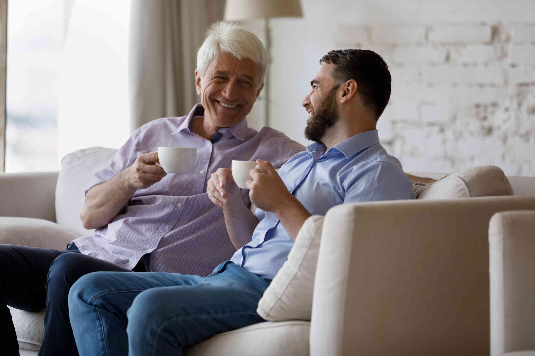 An elderly man and his adult son sit next to each other on the couch while holding while coffee cups and smiling at one another.